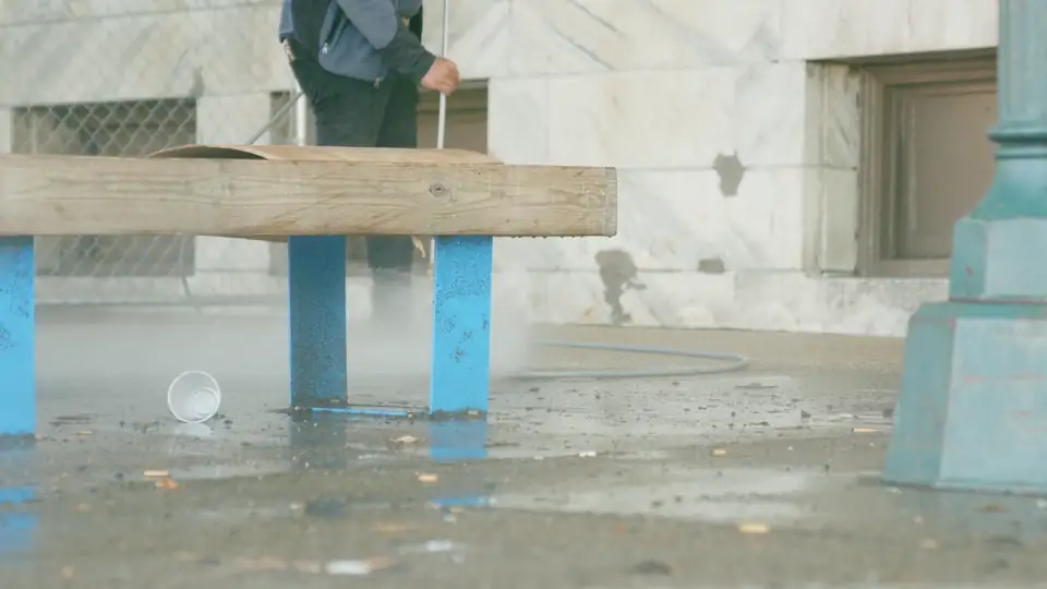 A pressure cleaner washing a paved outdoor area beside a bench